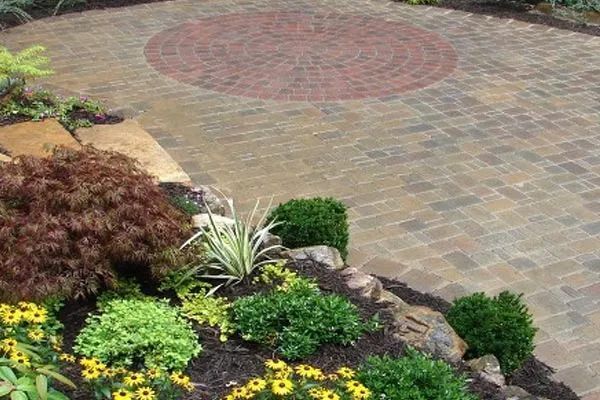 Brick patio with circular red brick accent, surrounded by landscaping with bushes and flowers.