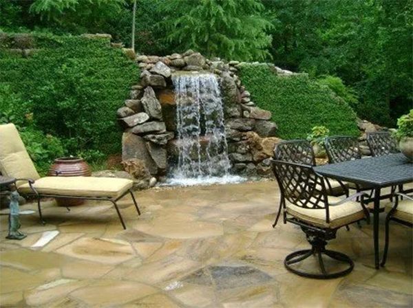 Patio with waterfall feature, stone pavers, outdoor dining set, lounge chair, surrounded by greenery.