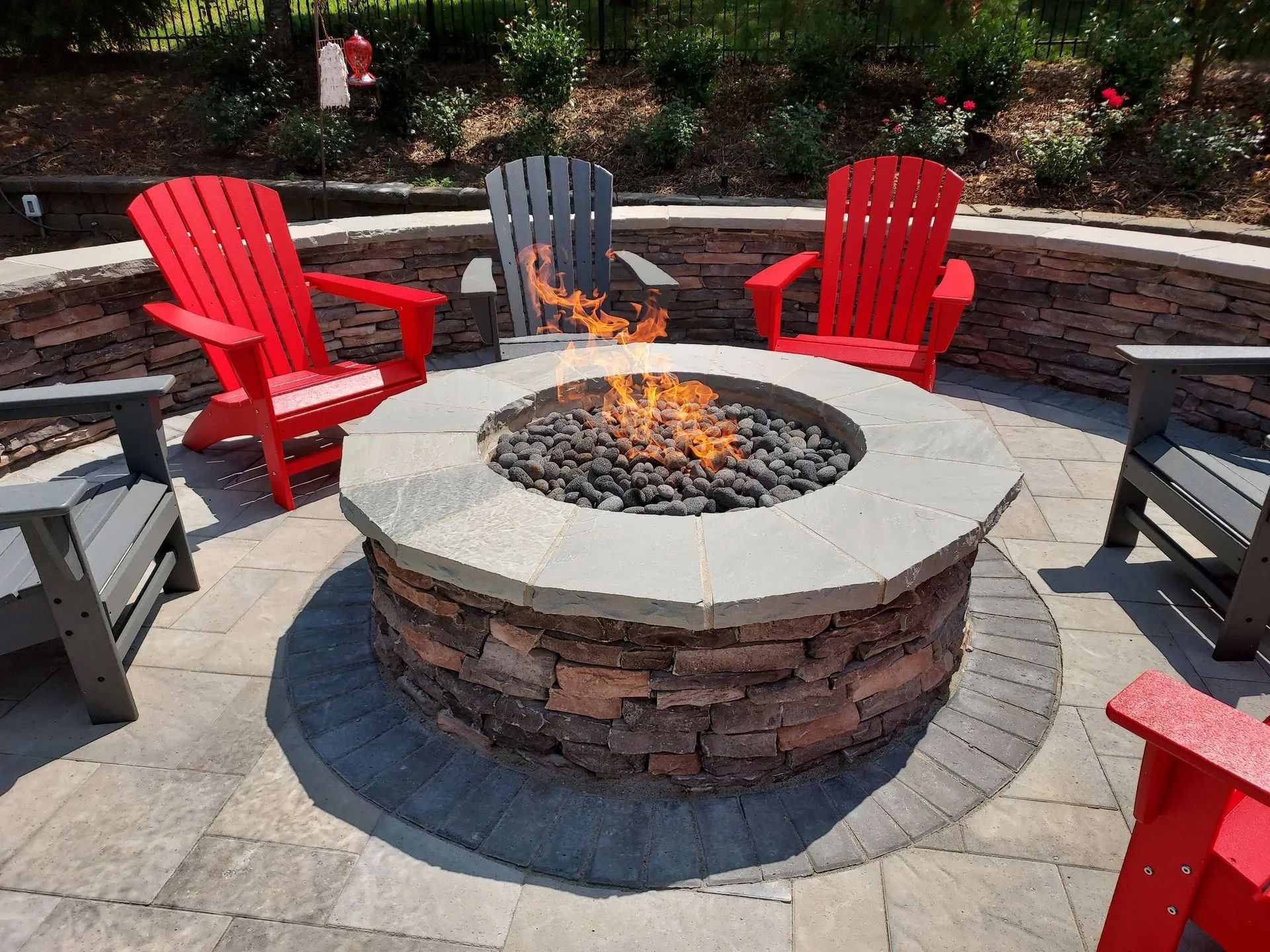 Fire pit with burning flames, surrounded by four red and gray Adirondack chairs, in a brick patio setting.