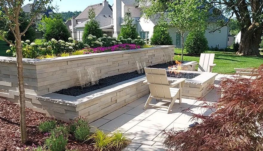 Stone wall fountain with black rocks, patio with fire pit and chairs, and landscaped yard.