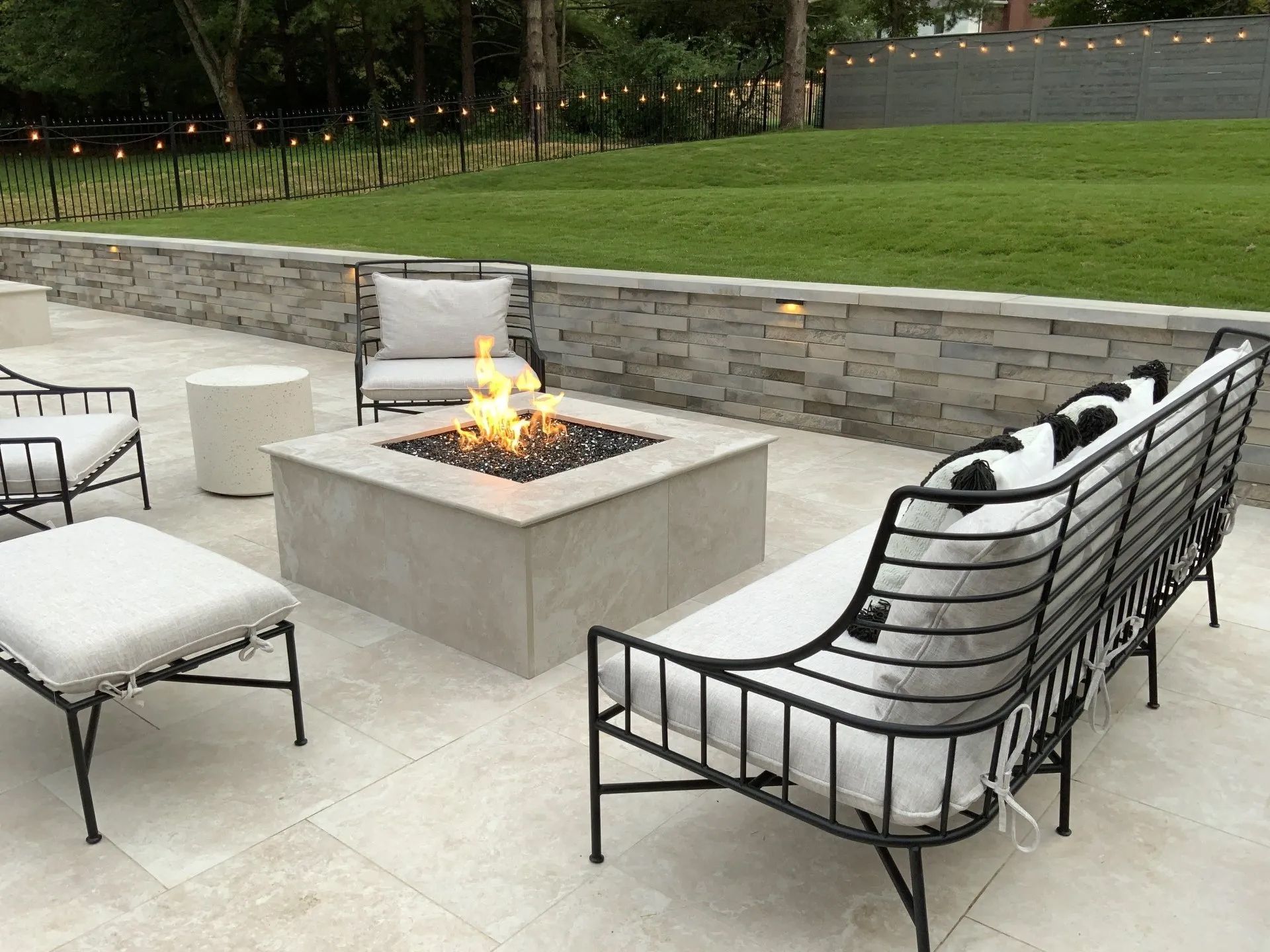 Outdoor patio with fire pit, seating, stone wall, and green lawn in background.