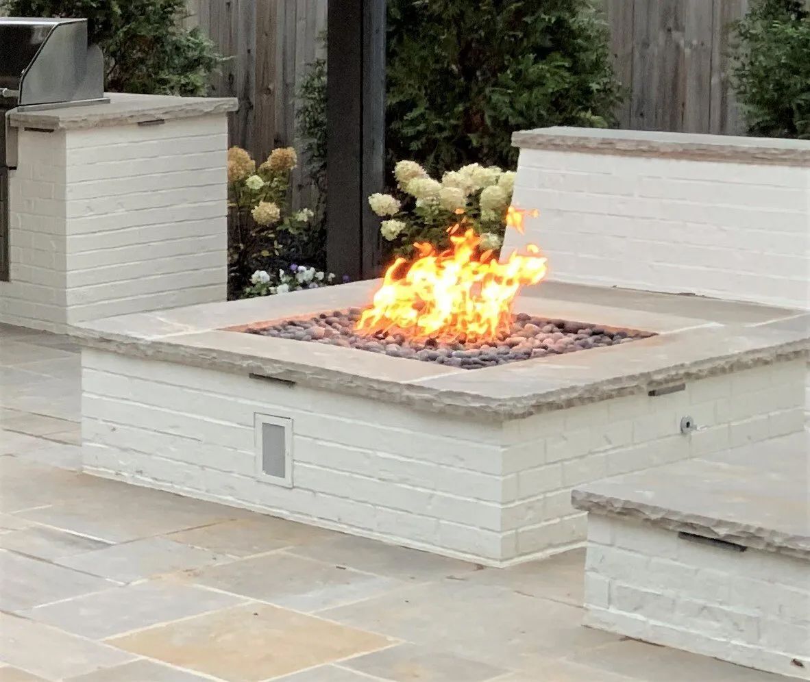 Stone fire pit with flames, surrounding seating on a patio. Grill station also in view.