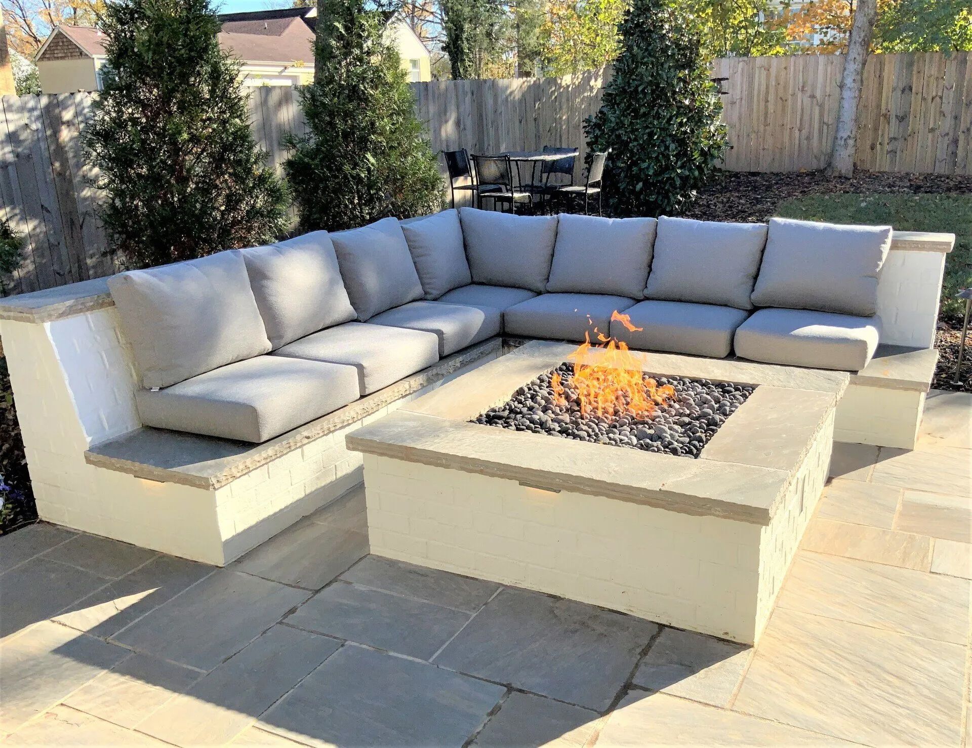 Outdoor sectional sofa with gray cushions, built-in around a fire pit with flames, on a stone patio.
