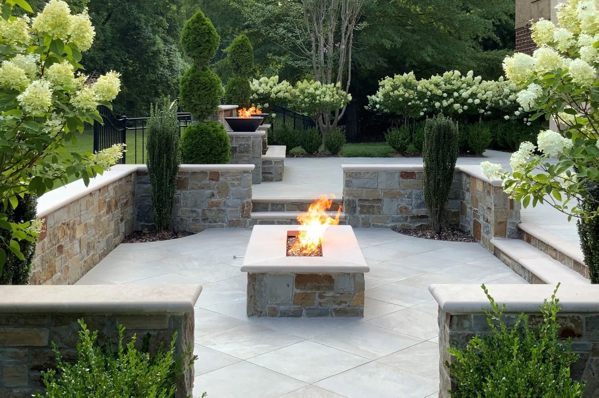 Stone patio with fire pit, raised walls, and landscaped garden with shrubs and trees.