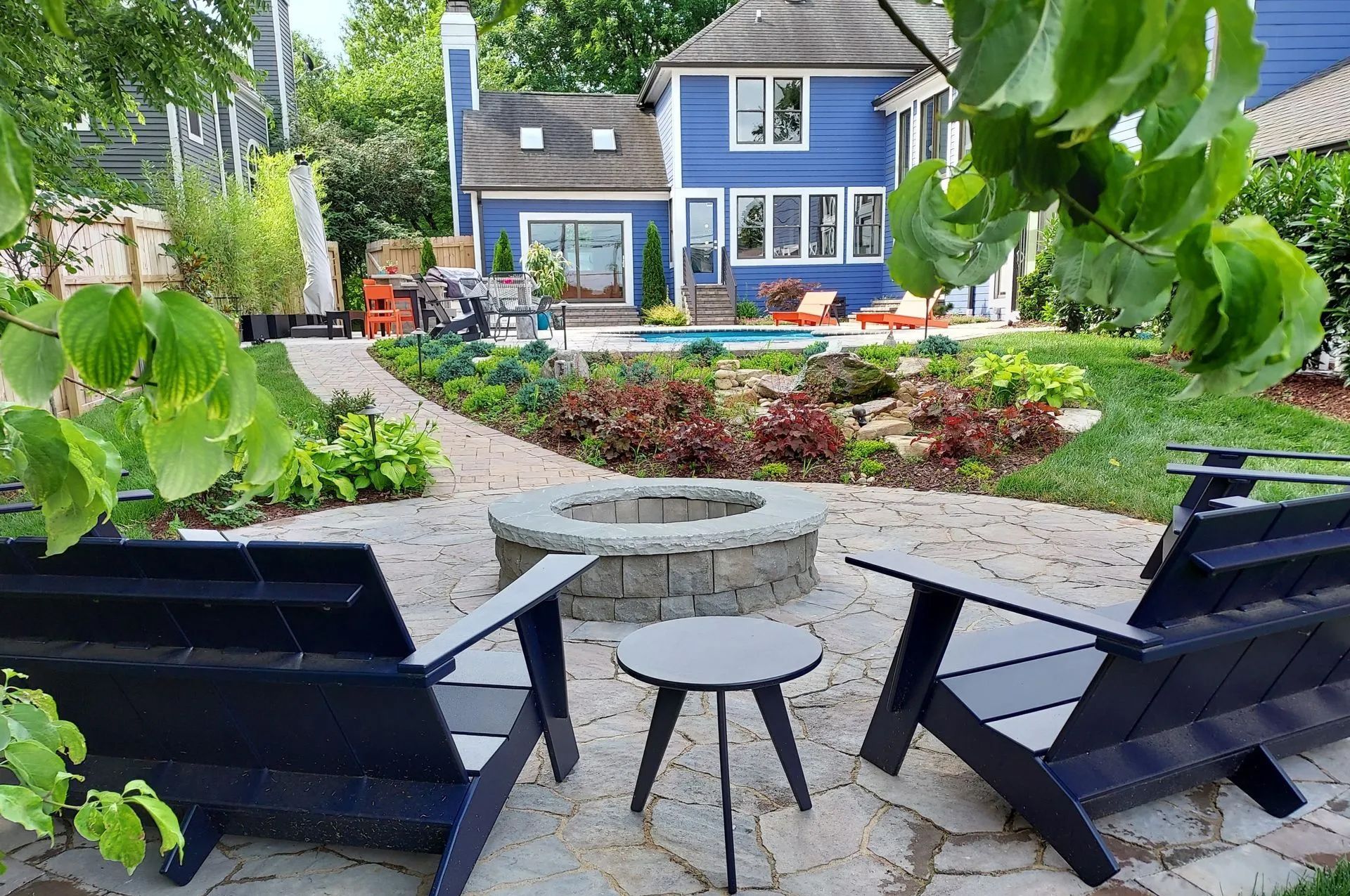 Backyard patio with fire pit, blue house, chairs, and garden path.