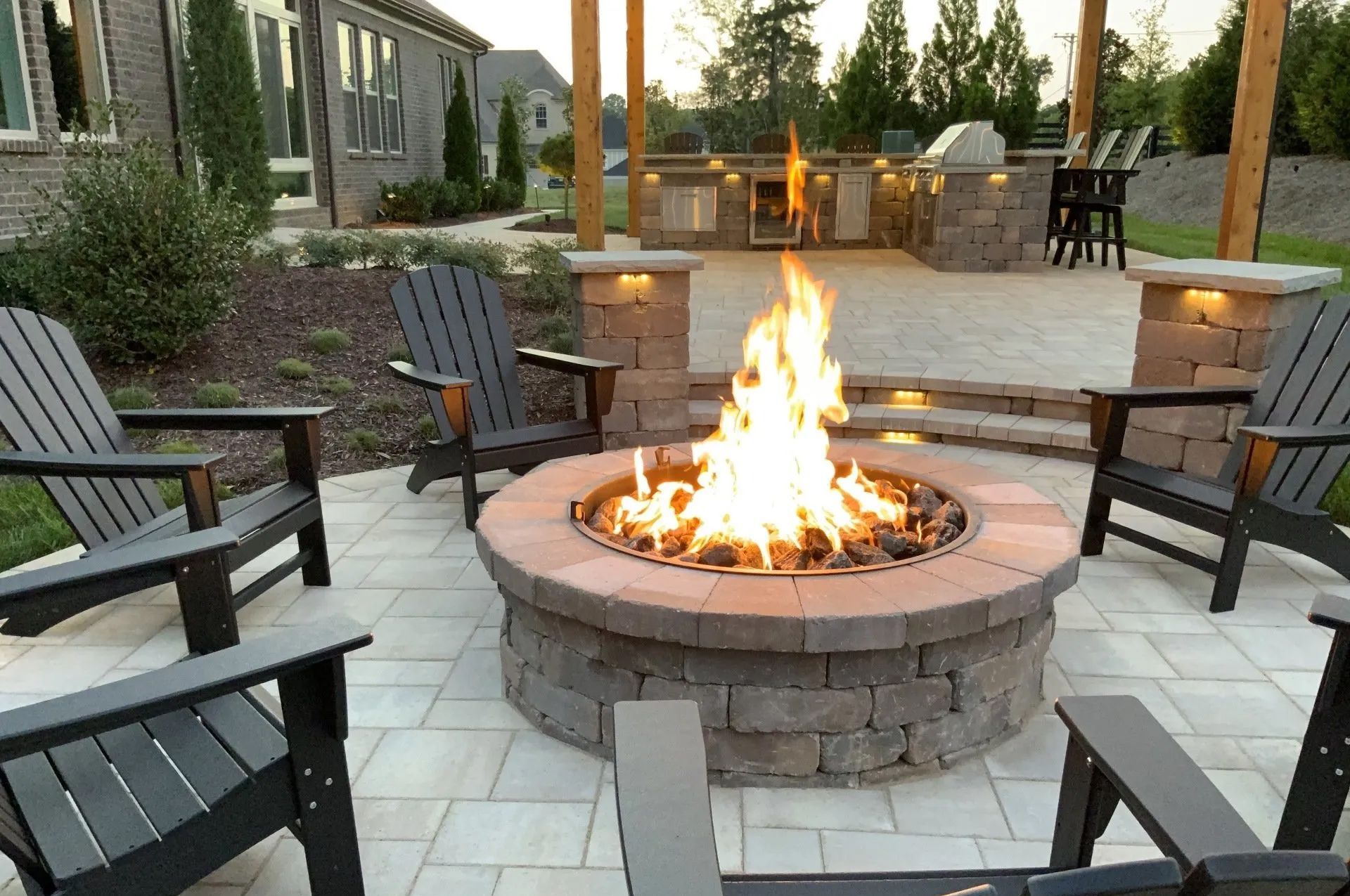 Stone fire pit with roaring flames, surrounded by Adirondack chairs on a paved patio, outdoor kitchen in the background.
