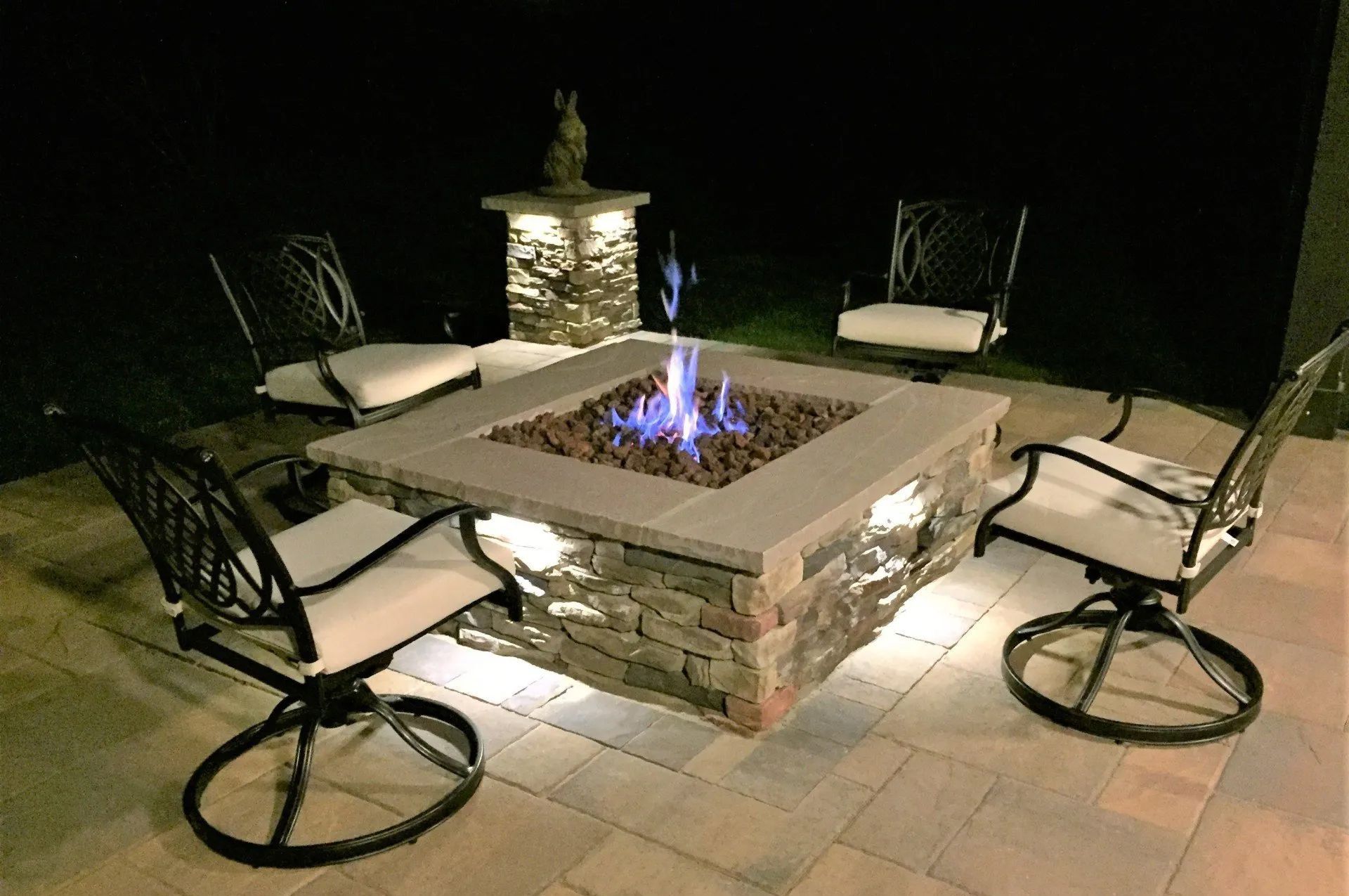 Fire pit surrounded by four chairs on a patio at night with lit flames and ambient lighting.