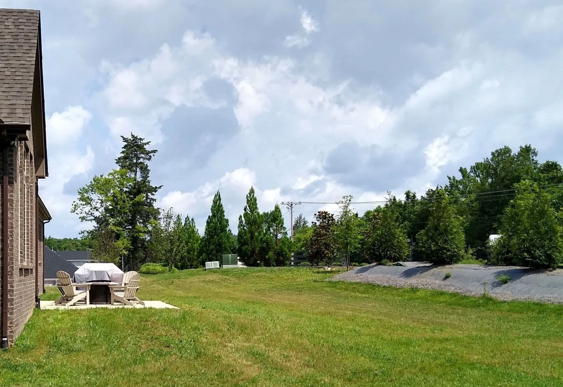 Grassy backyard with trees, a brick house, and a stone fire pit under a cloudy sky.