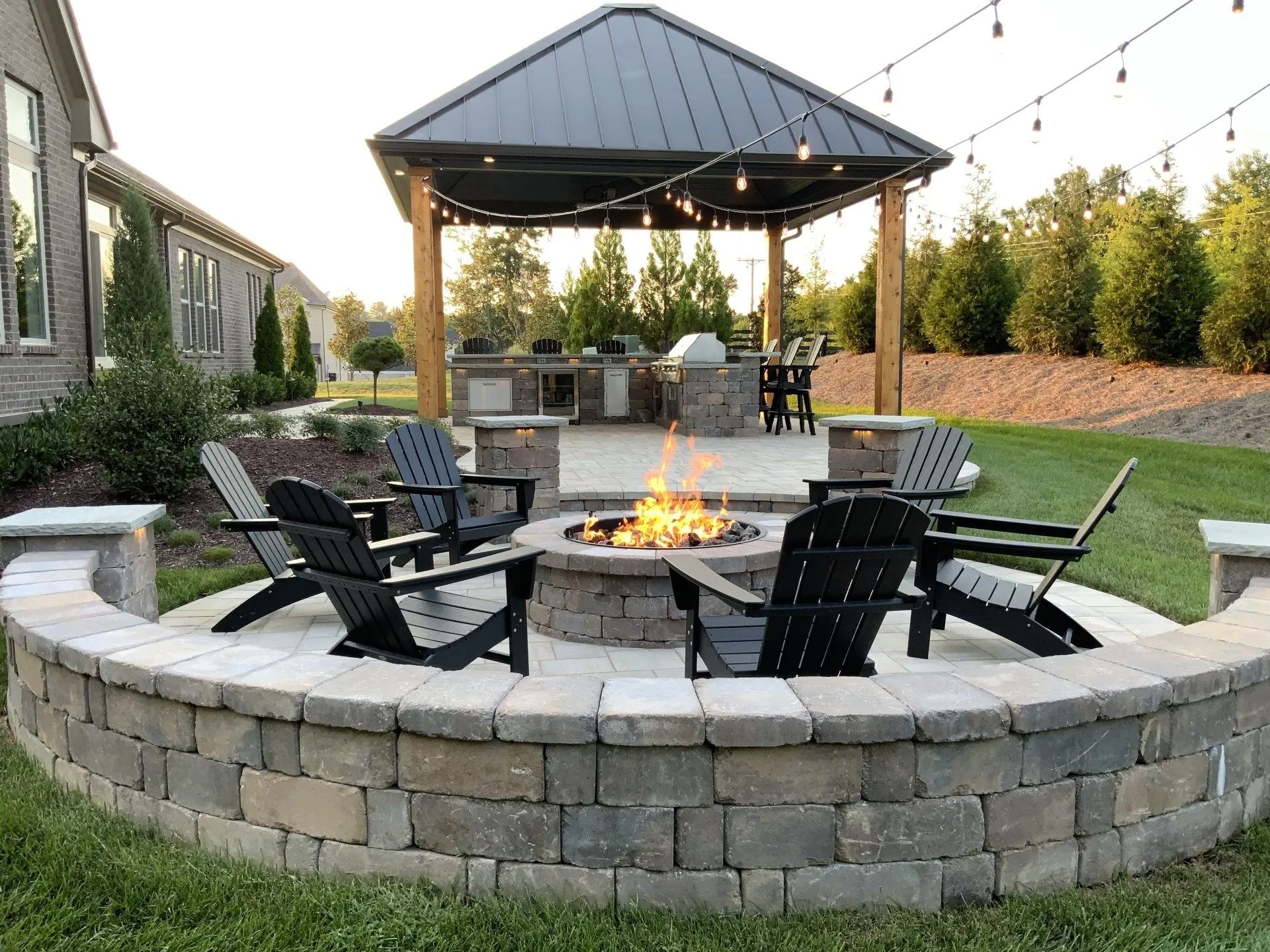 Fire pit with Adirondack chairs on a stone patio, outdoor kitchen and gazebo in the background.