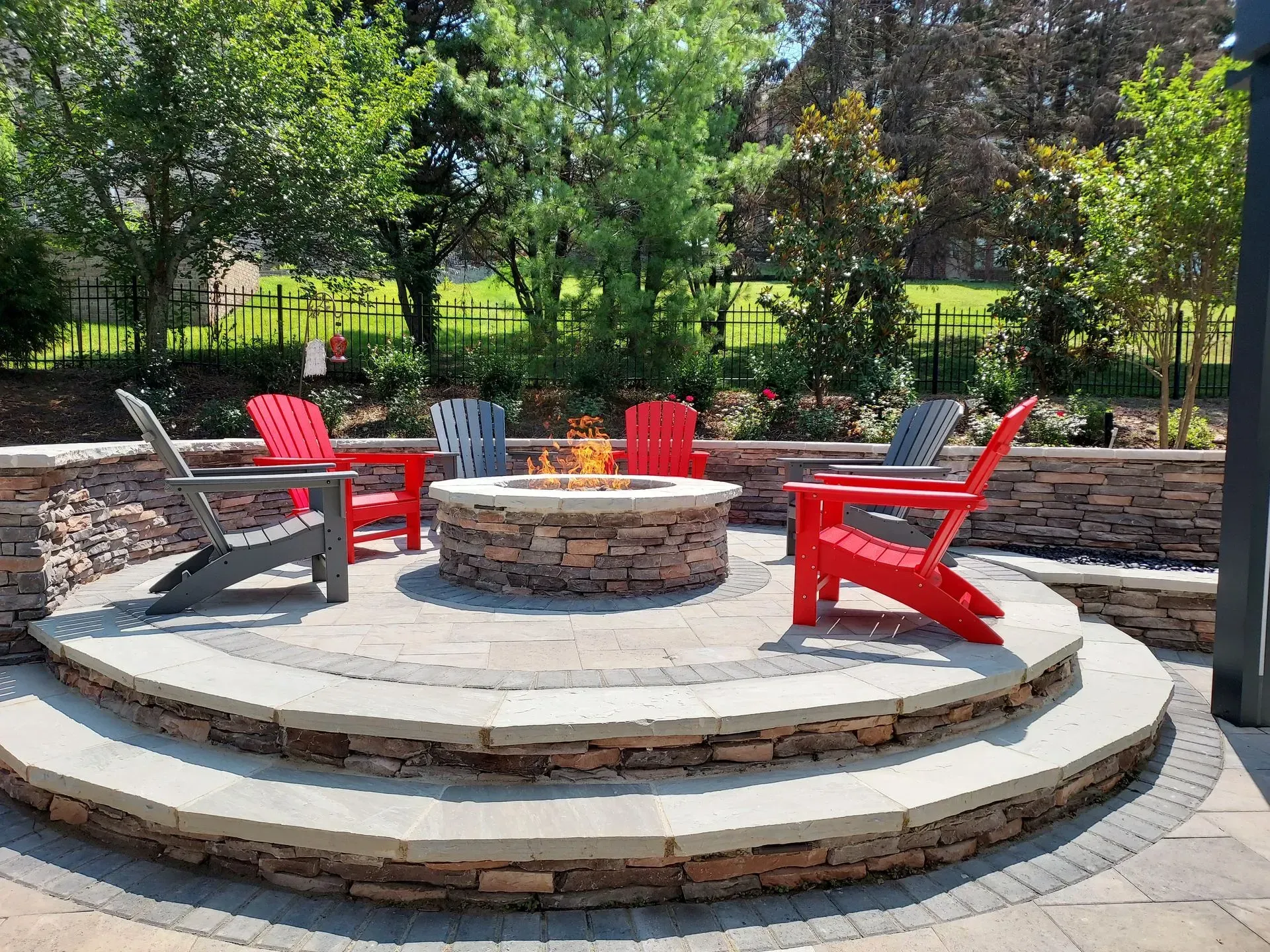 Fire pit with red and gray Adirondack chairs on a stone patio in a backyard with trees.