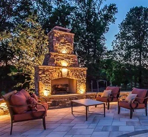 Outdoor stone fireplace with chairs and a table on a patio, lit at dusk.