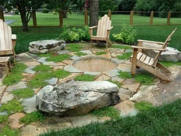 Fire pit area with stone pavers, wooden chairs, and large rocks in a grassy yard.