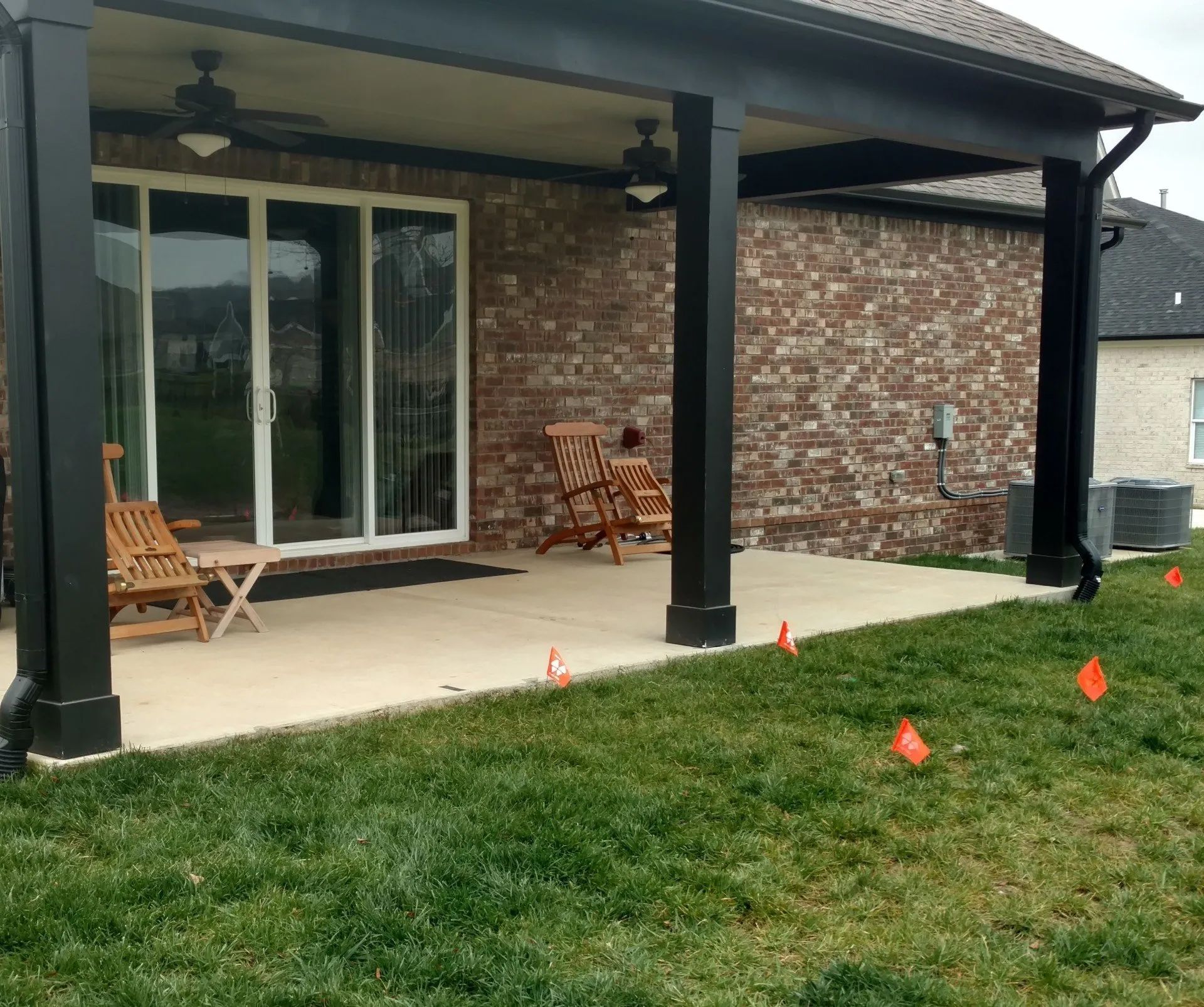 Covered patio with black columns, brick wall, and sliding glass door; two chairs sit on the concrete patio.
