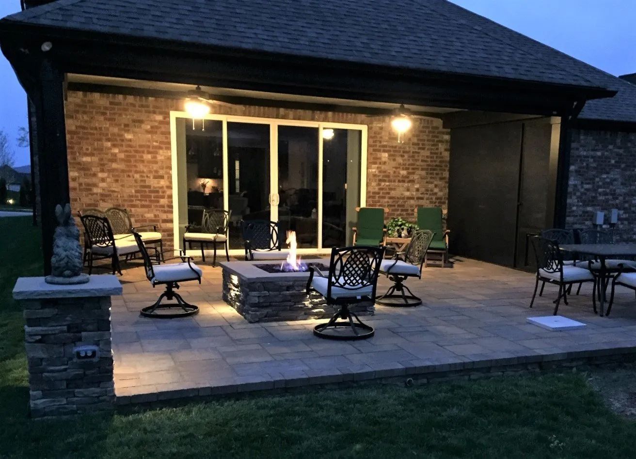 Patio with fire pit and seating, lit by overhead lights and house lighting at dusk.