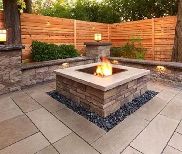 Outdoor fire pit surrounded by stone patio, wooden fence, and landscape lighting.