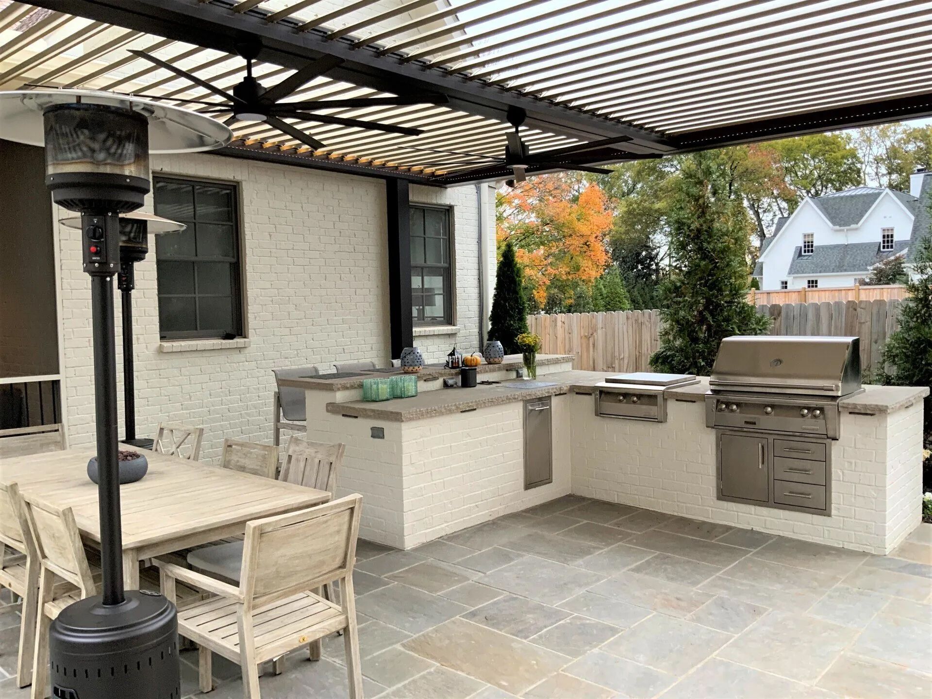 Outdoor kitchen with grill, dining table, and heaters under a pergola.