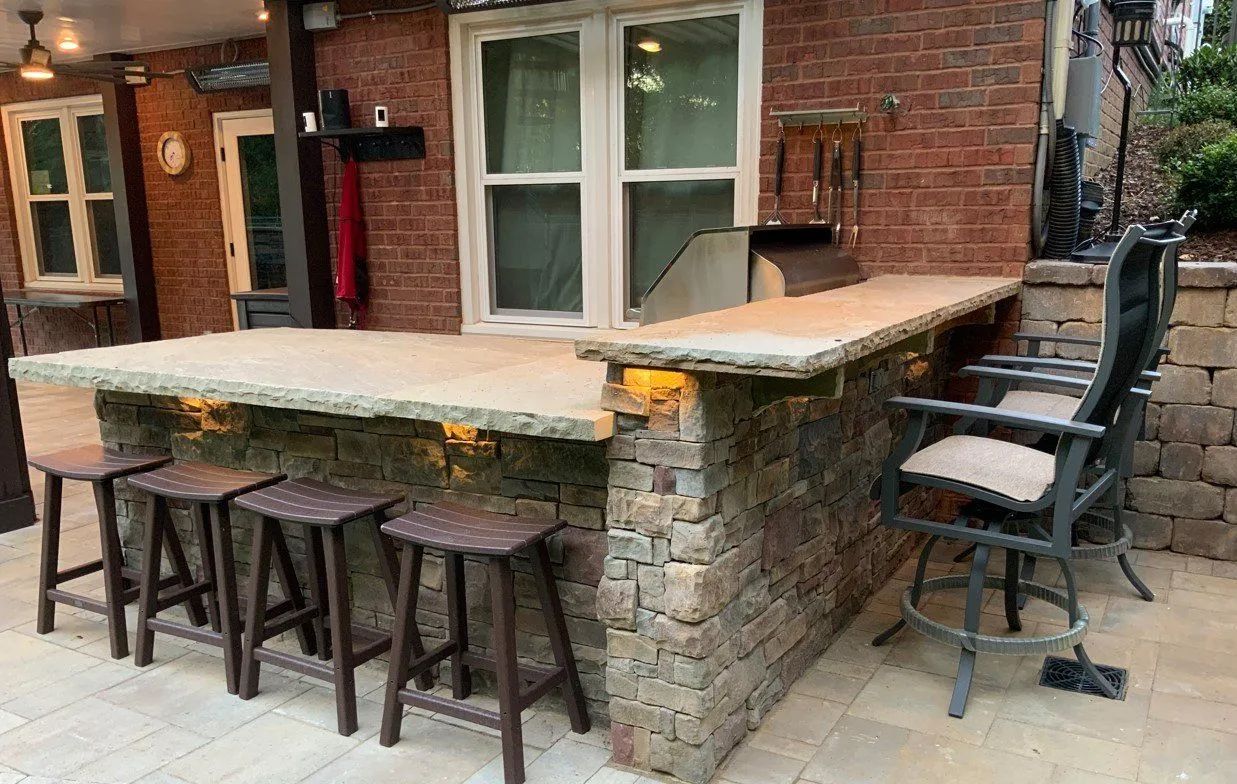 Outdoor kitchen with stone bar, stools, and built-in grill next to a window.