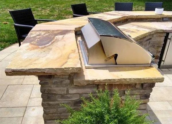 Outdoor kitchen island with built-in grill, stone countertop, brick base, and seating area in a backyard setting.
