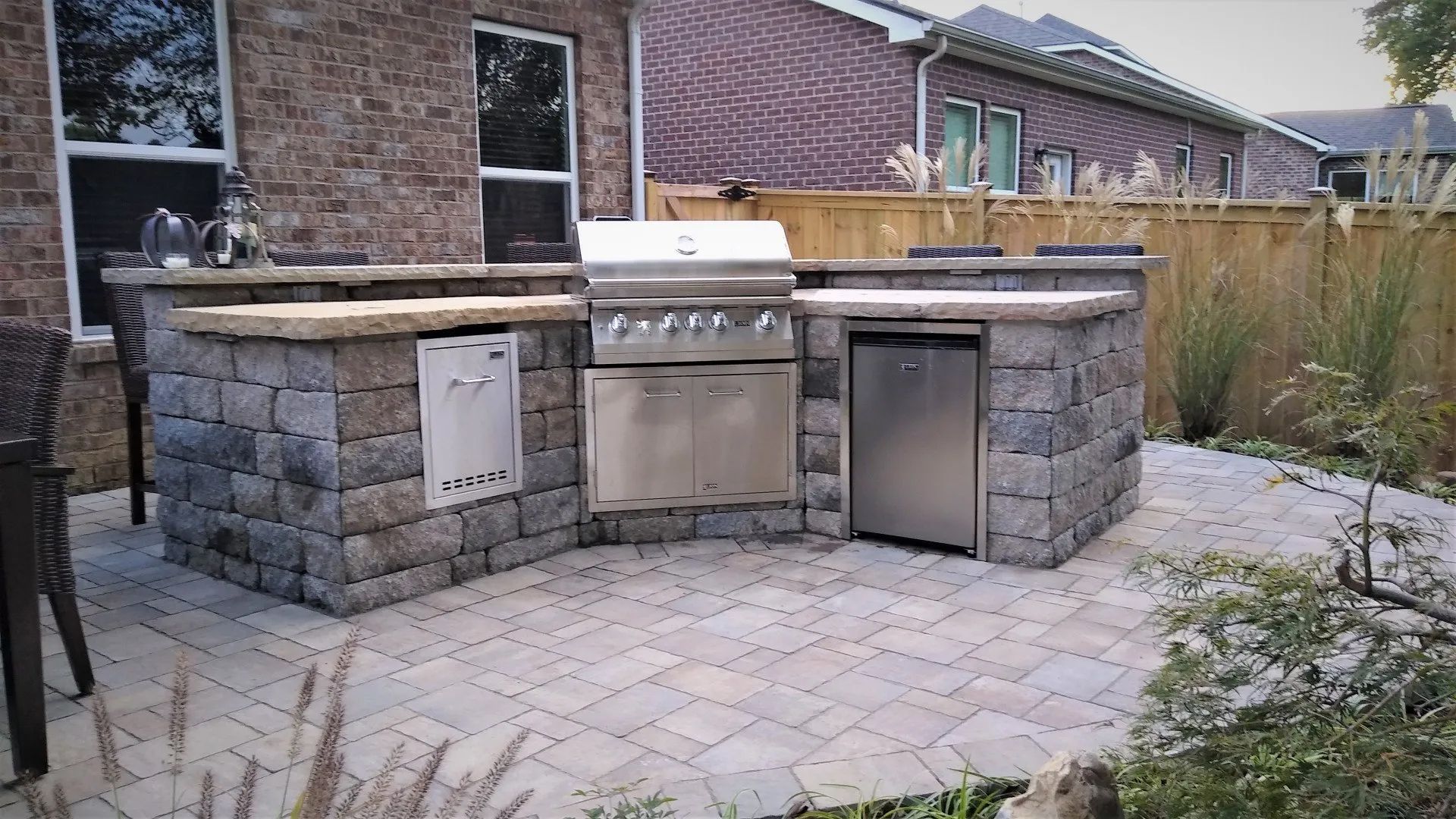 Outdoor kitchen with grill, refrigerator, and stone countertops, on a brick patio.