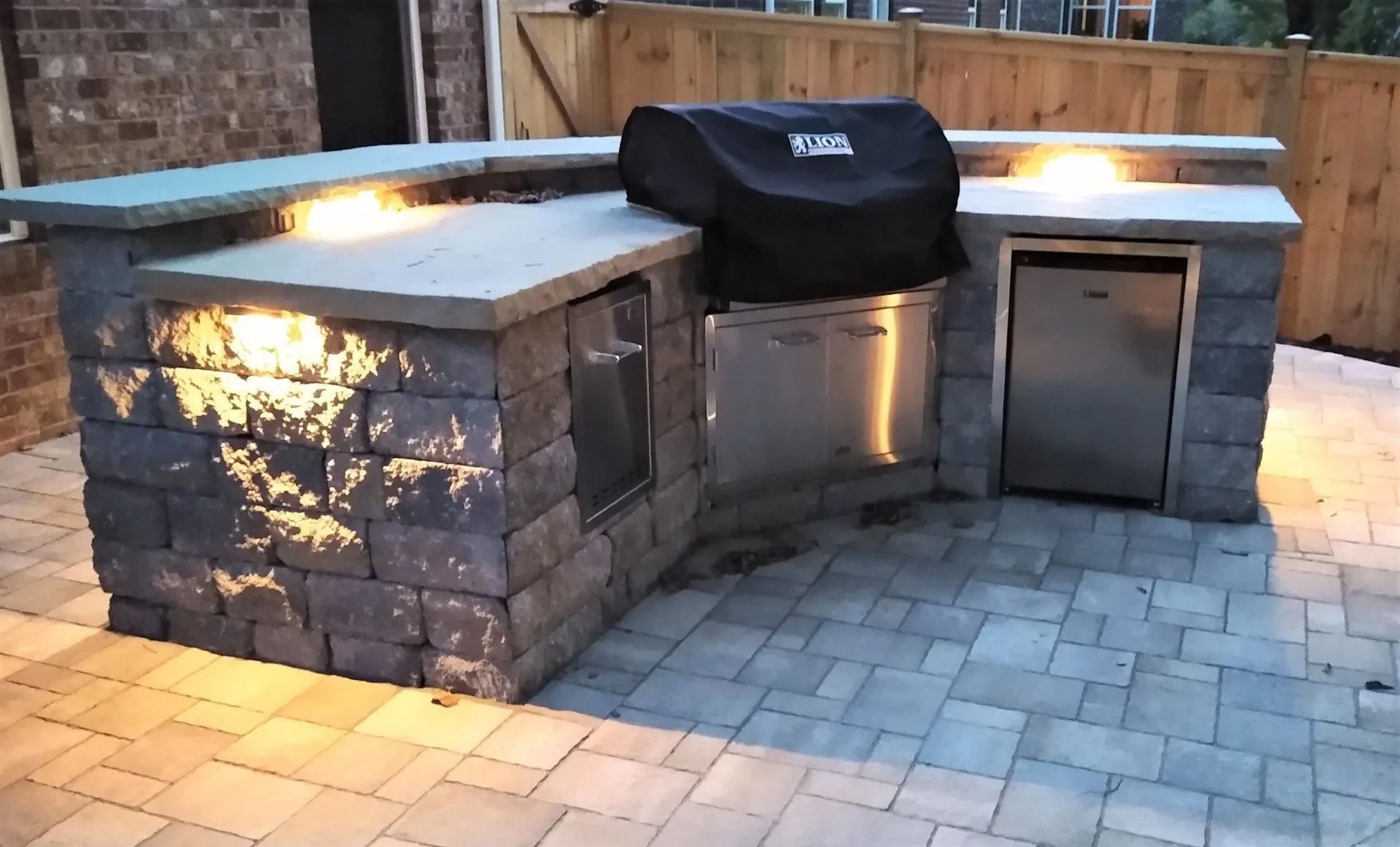 Outdoor kitchen built with stone blocks, with grill, refrigerator, and countertop with lighting.