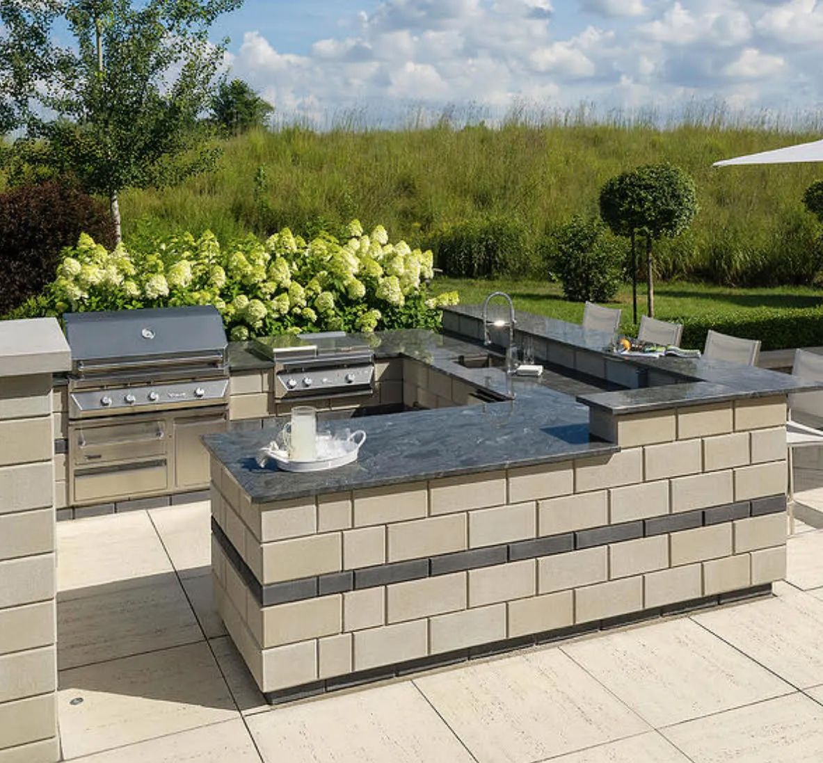 Outdoor kitchen island with grill, countertop, and seating area on a patio, surrounded by greenery.