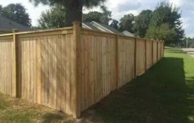 Wooden fence in a yard, trees and houses in the background, green grass.