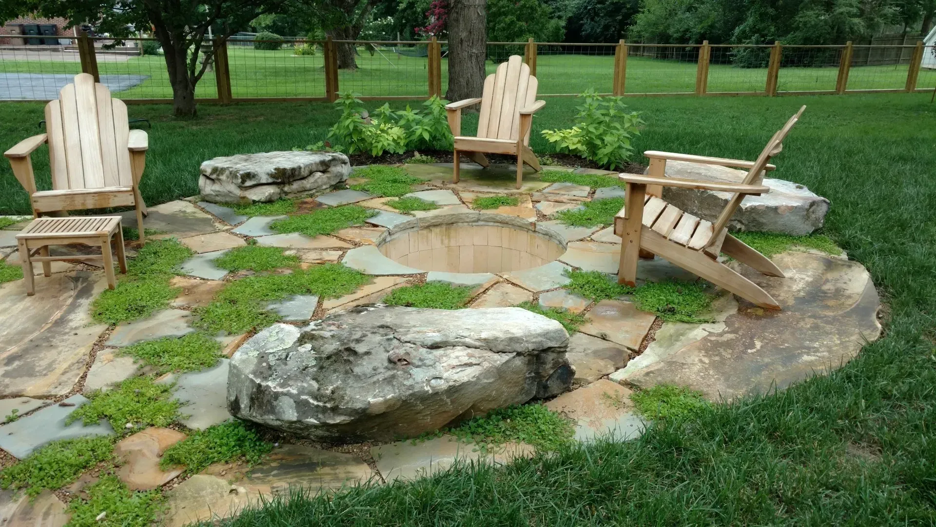 Fire pit area with wood chairs on a stone patio surrounded by grass and a wooden fence.