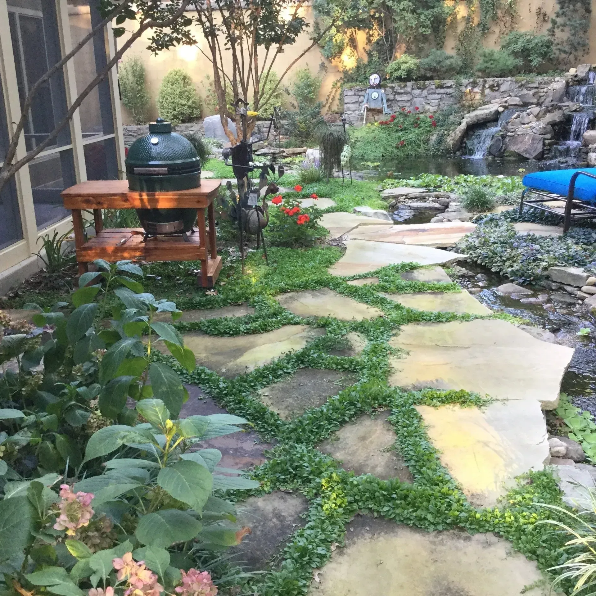 Stone path through lush garden with waterfall, grill, and greenery.