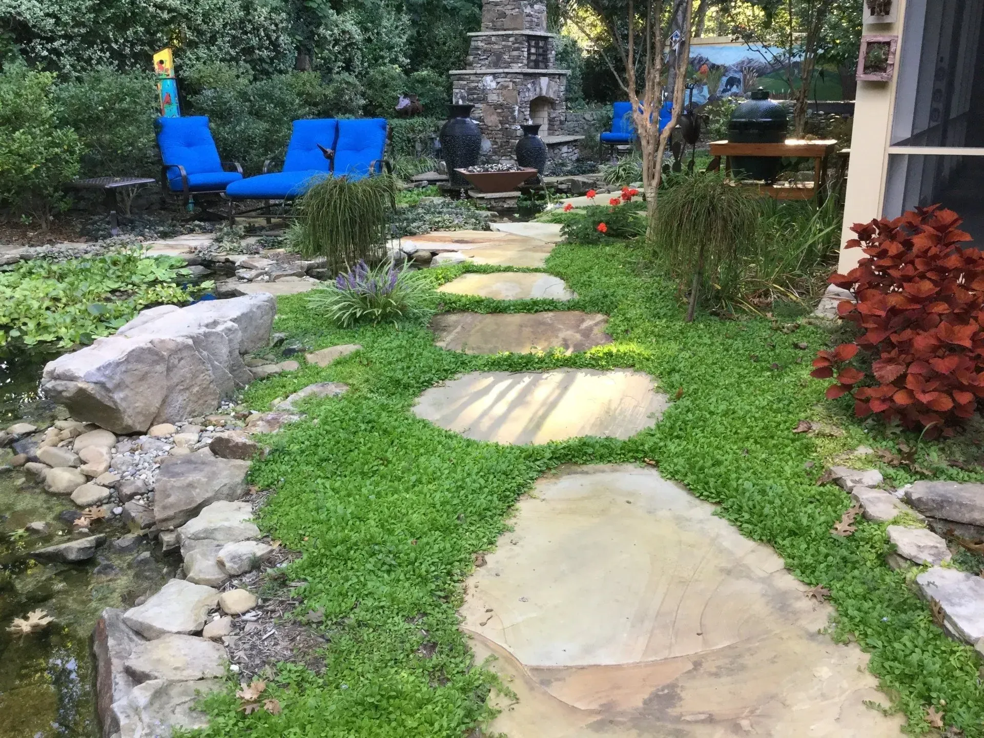 Stone path through a garden, leading to a seating area with blue chairs. Green ground cover, pond nearby.