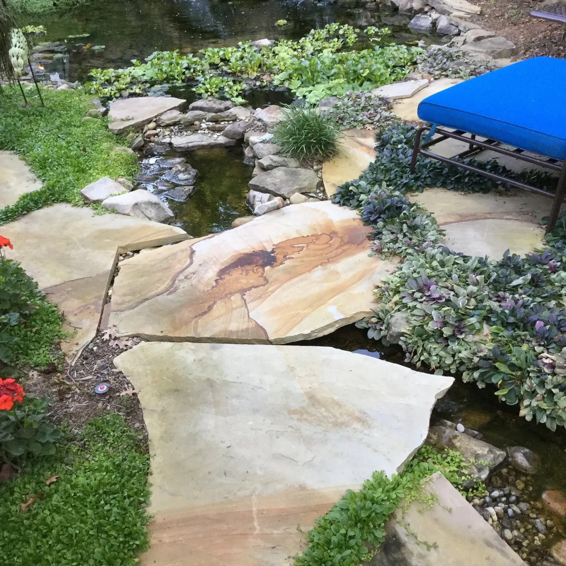 Flagstone path leads over a small stream in a garden. Blue chair sits nearby.
