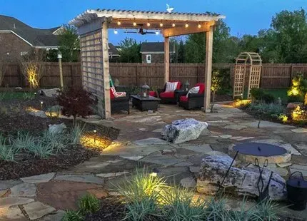 Backyard patio with wooden pergola, seating, fire pit, and landscaping at dusk.