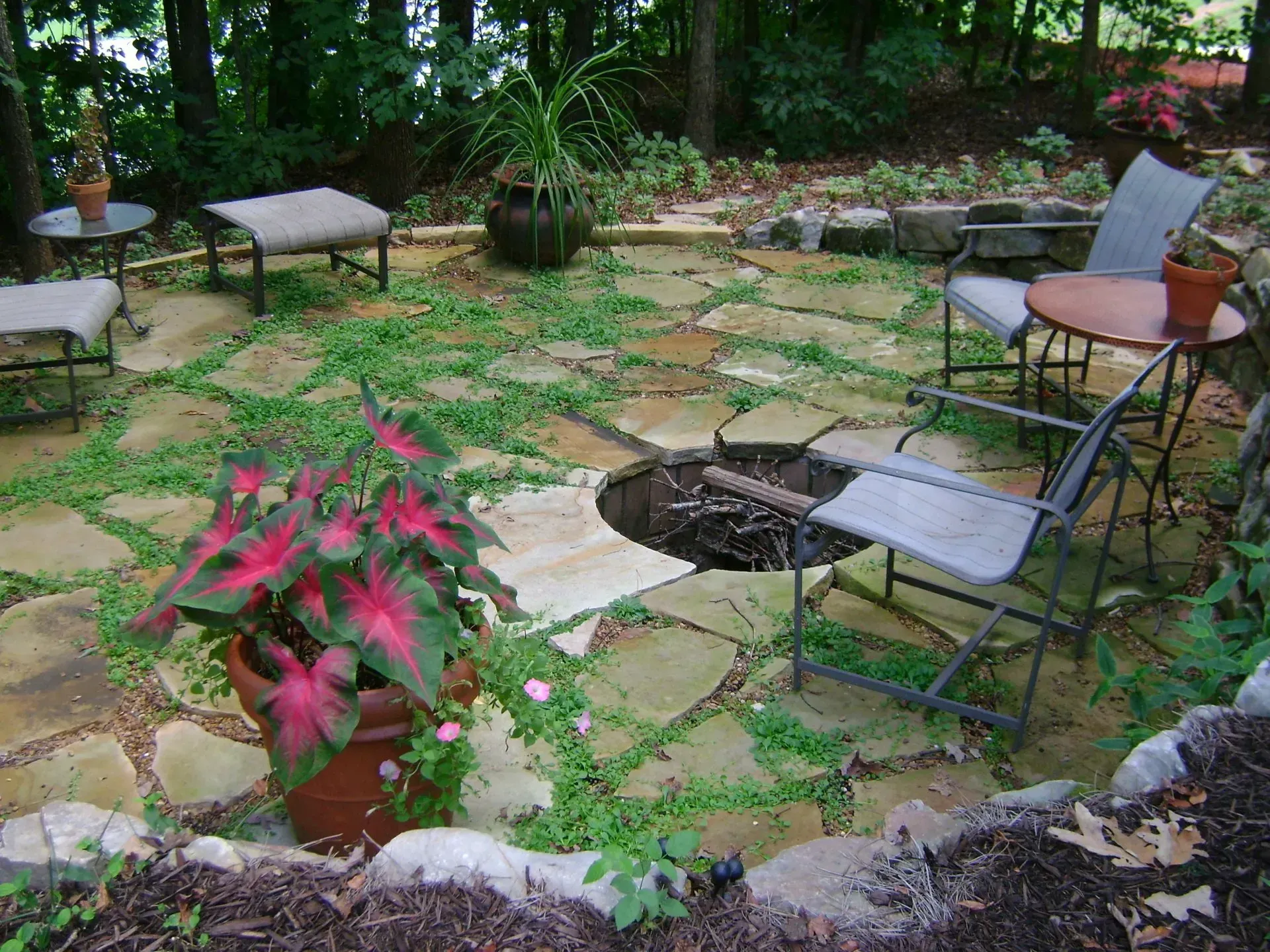 Stone patio with fire pit, seating, and potted plants, surrounded by greenery and trees.