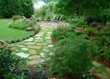 Stone path through a lush garden leads to patio furniture. Green foliage, grass, and trees surround.