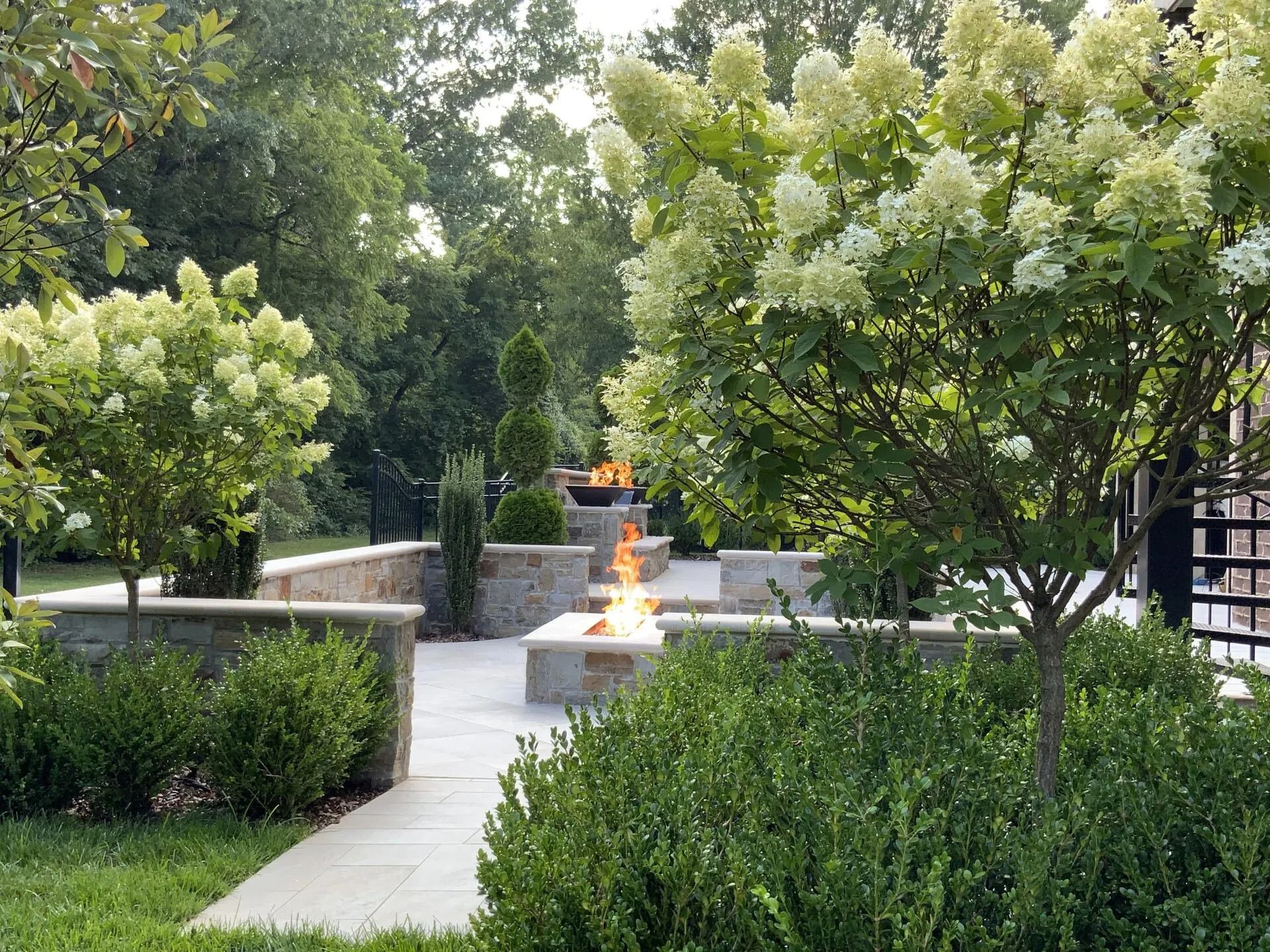 Stone patio with fire pits surrounded by manicured greenery and trees.