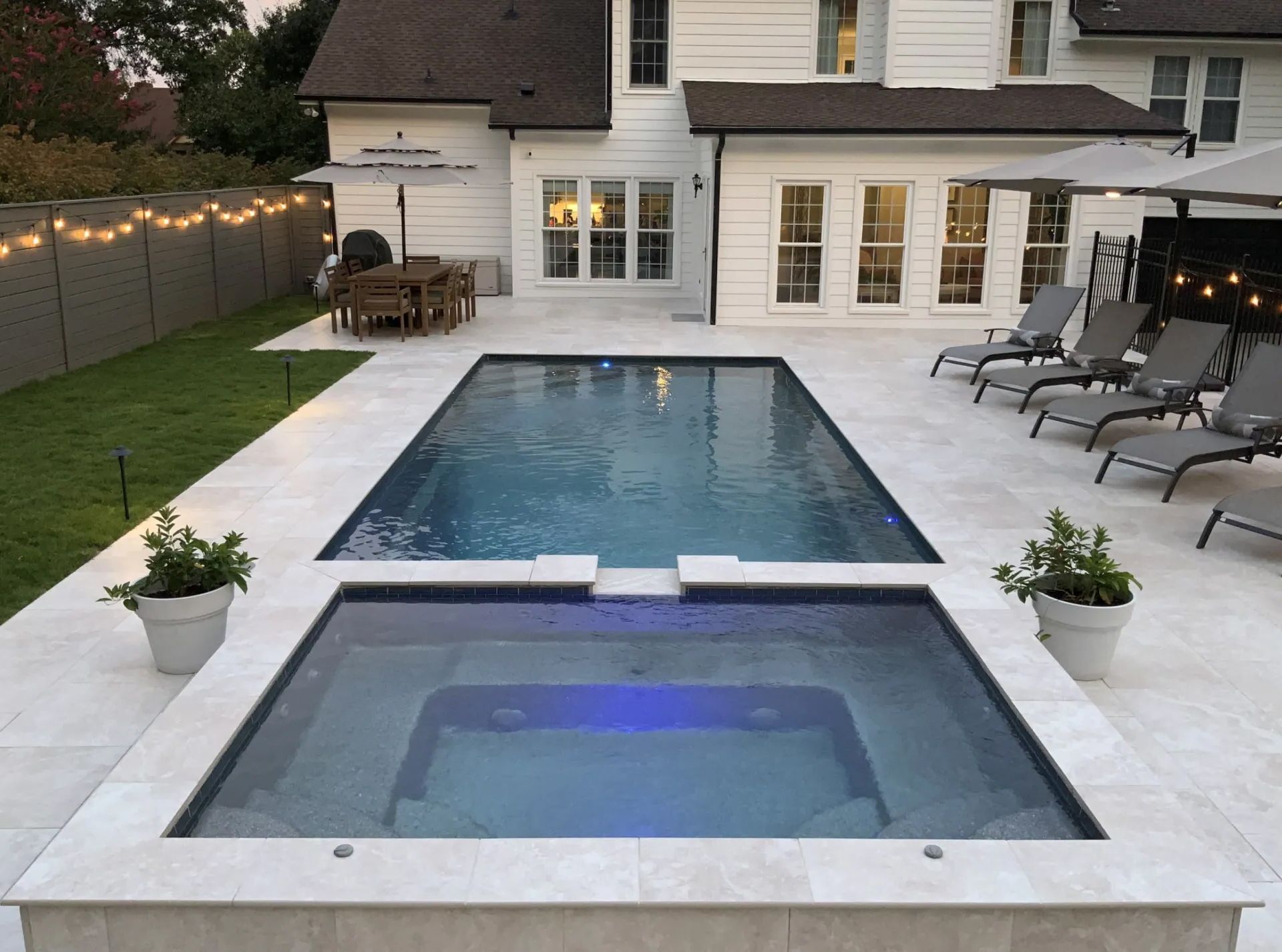 Backyard with pool and spa, white house, lounge chairs, patio, and string lights.