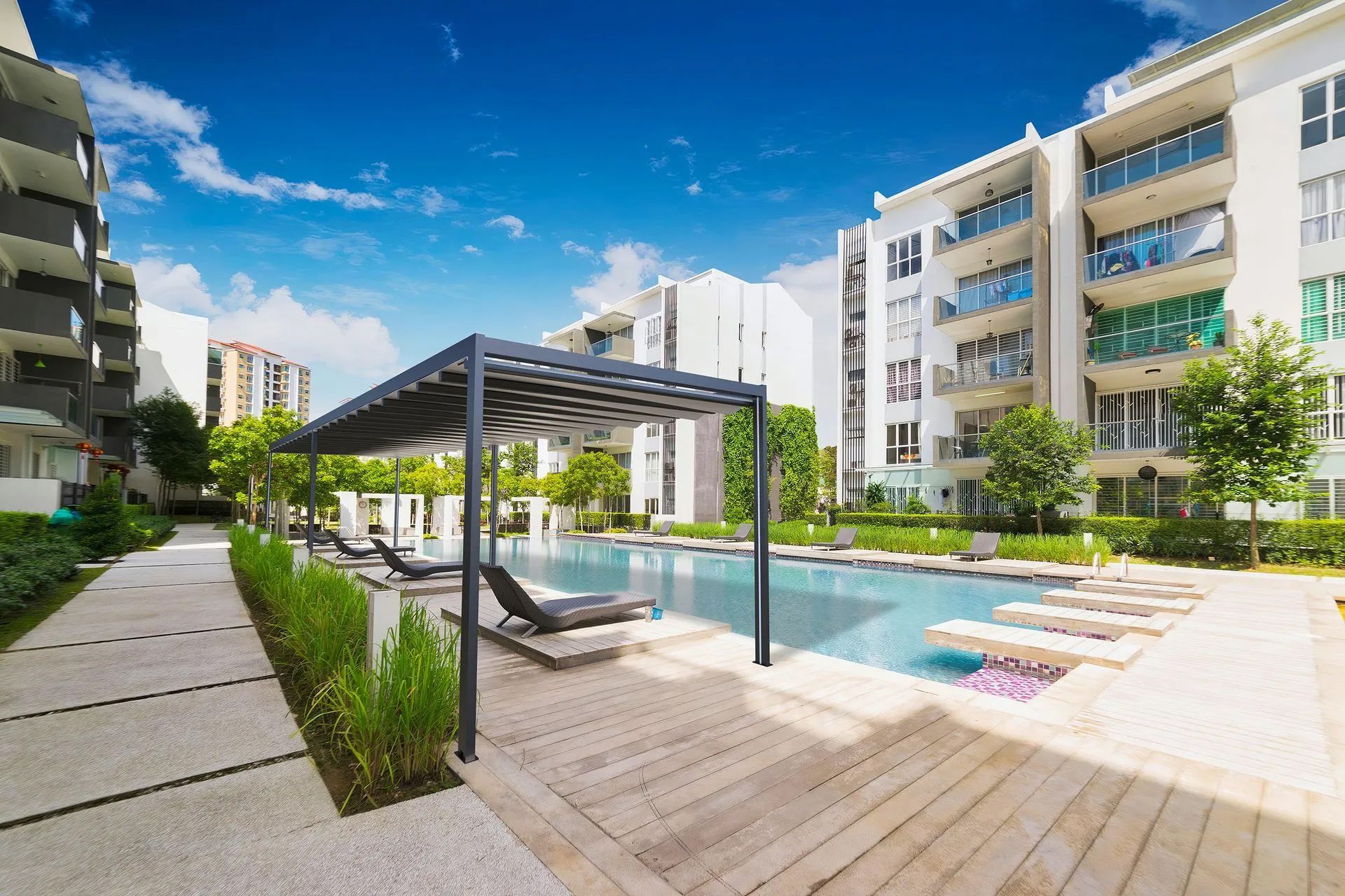 Pool area with lounge chairs, pergola, and apartment buildings under a blue sky.