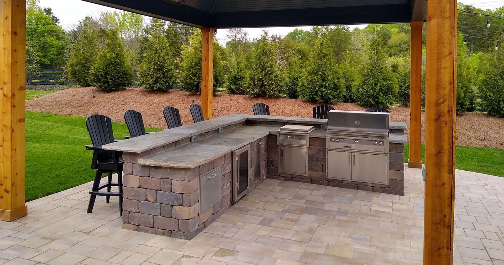 Outdoor kitchen with stone counters, built-in grill, and bar seating under a wooden pergola.