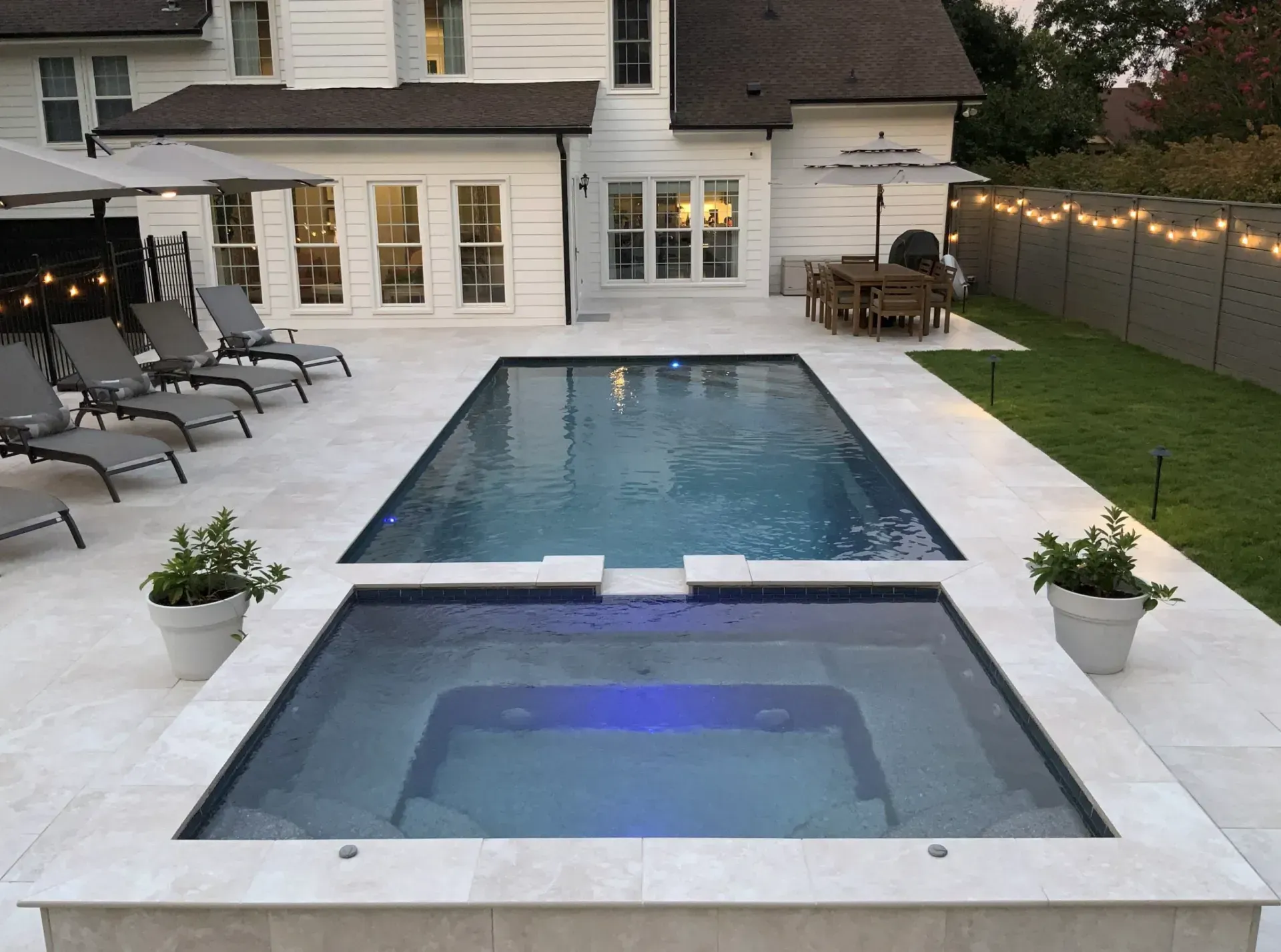 Backyard pool and spa with lounge chairs and dining area next to a white house.