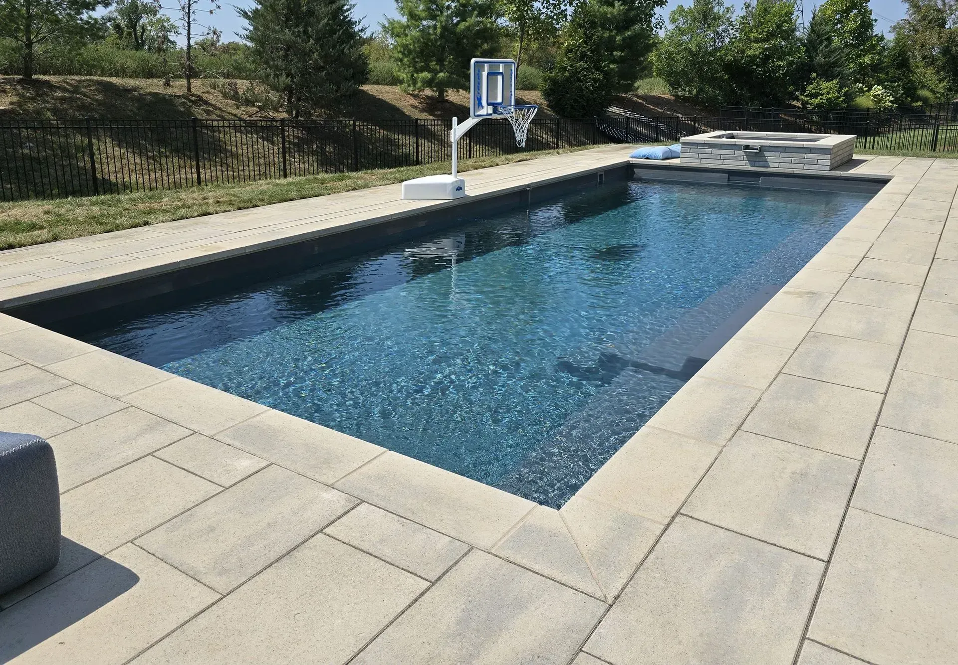 Rectangular pool with blue water, concrete patio, basketball hoop. Sunny outdoor setting.