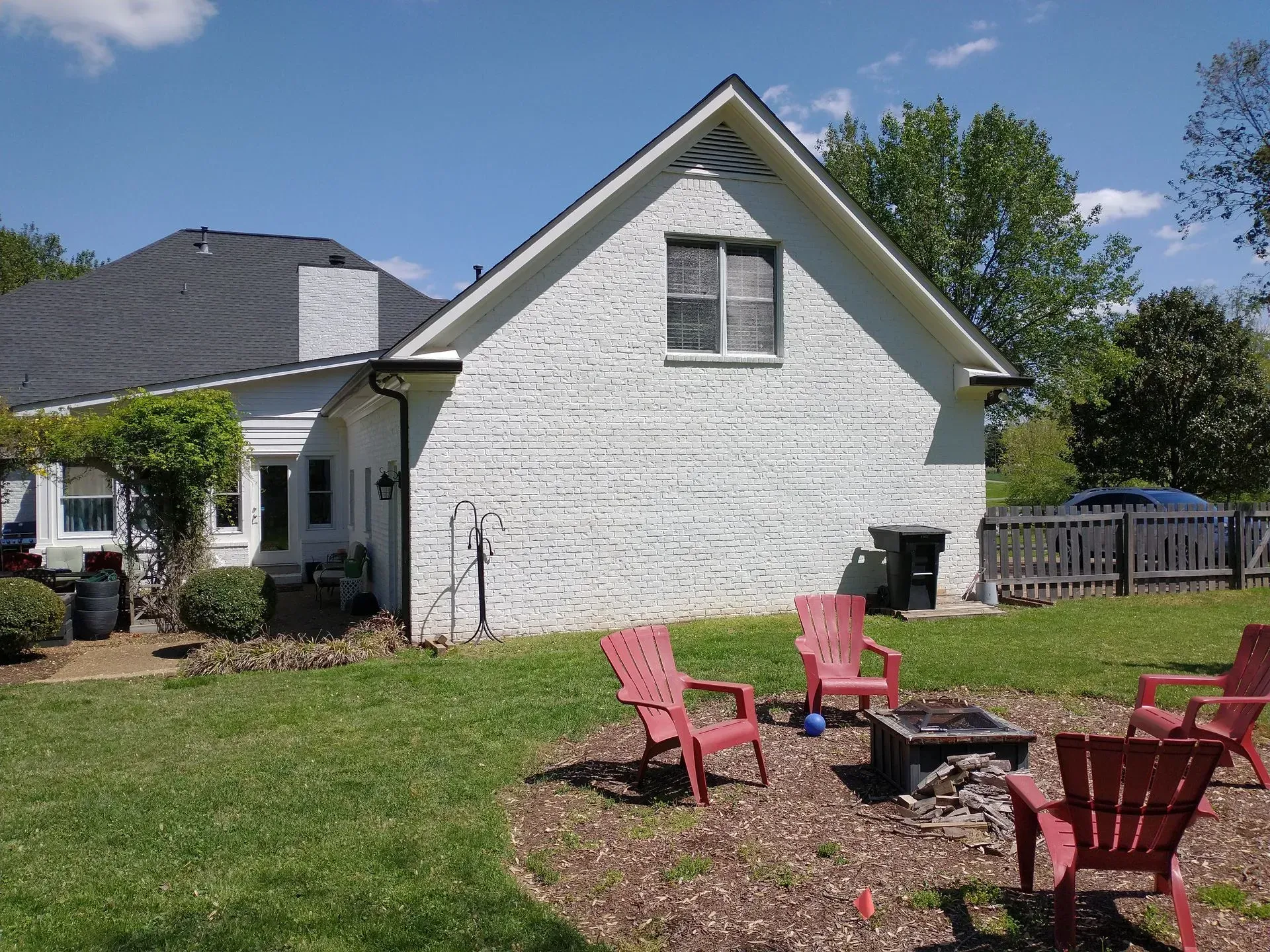 Backyard scene with red chairs around a fire pit and a white brick house under a blue sky.