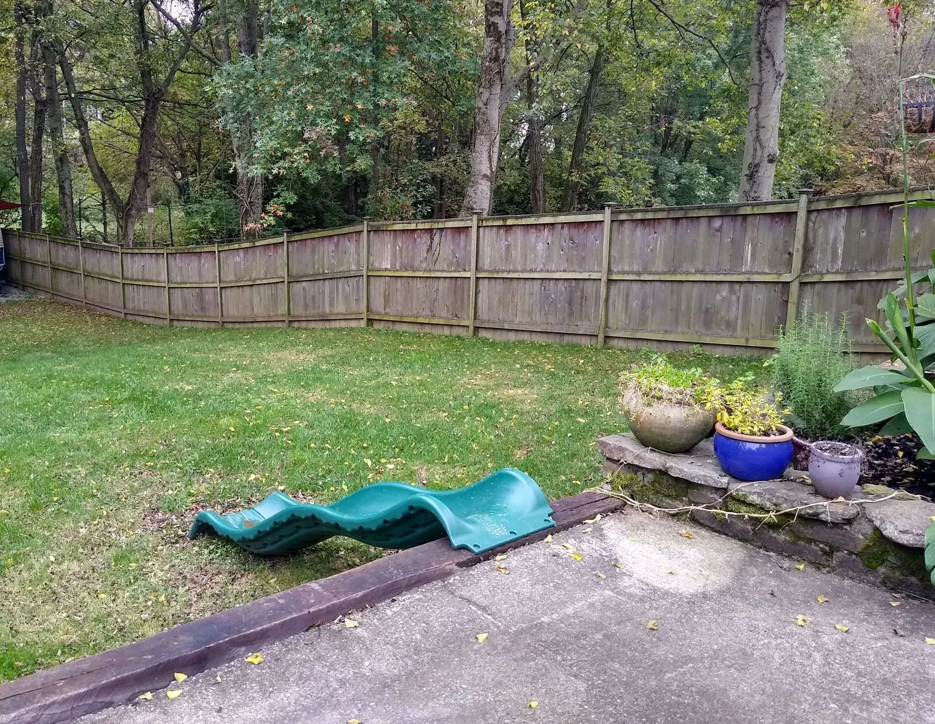 Green backyard with a wooden fence, grass, a slide, and potted plants.
