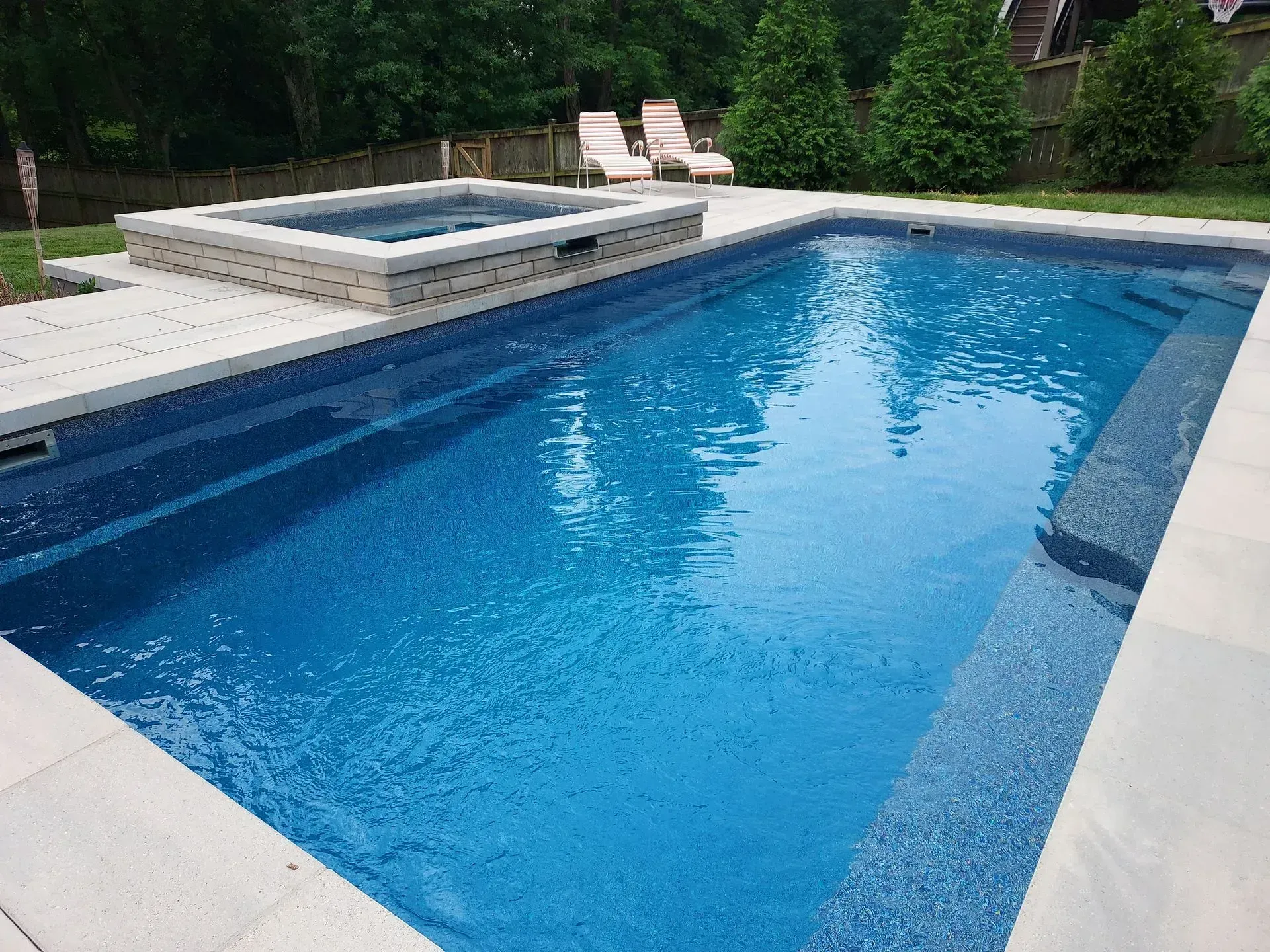 Swimming pool with built-in spa. Blue water, light stone patio, two lounge chairs in background.