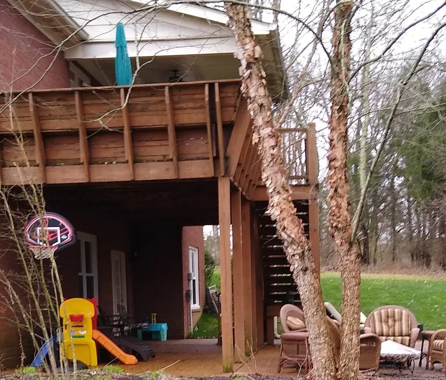 Wooden deck with stairs, basketball hoop, and children's play set beneath. Brown siding, green grass.