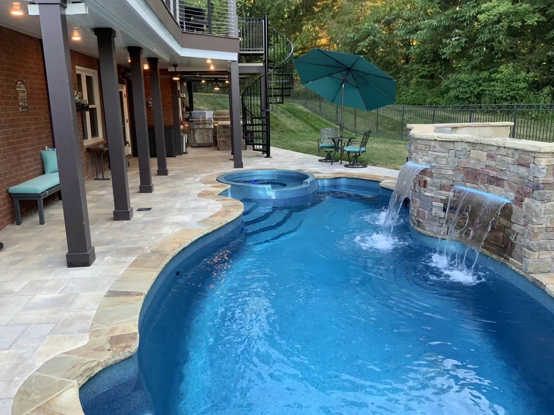 Pool with a waterfall, lounge area, and a spiral staircase leading to an outdoor kitchen and a patio.