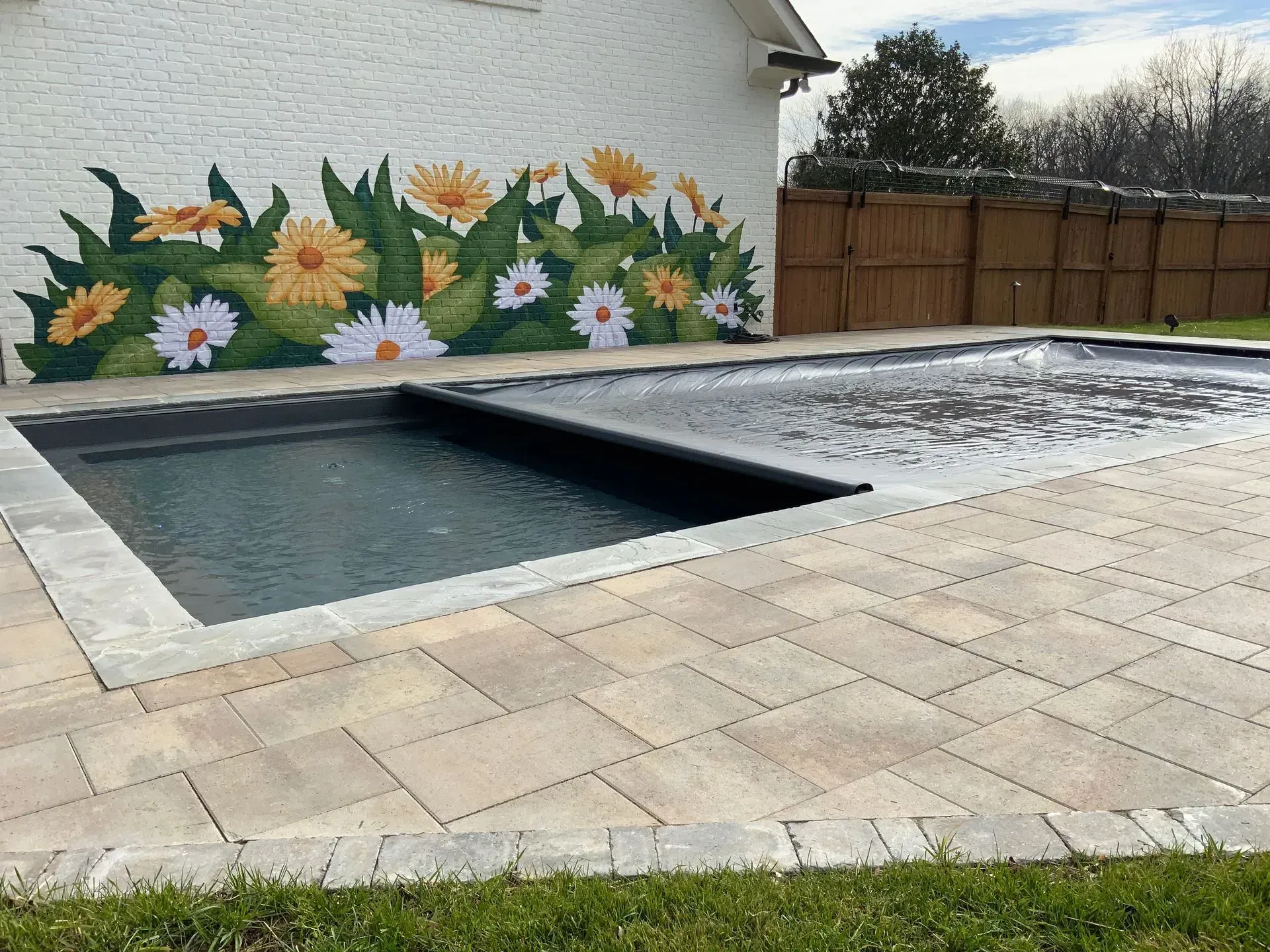 Rectangular pool with cover next to a brick building with a daisy mural, on a stone patio.