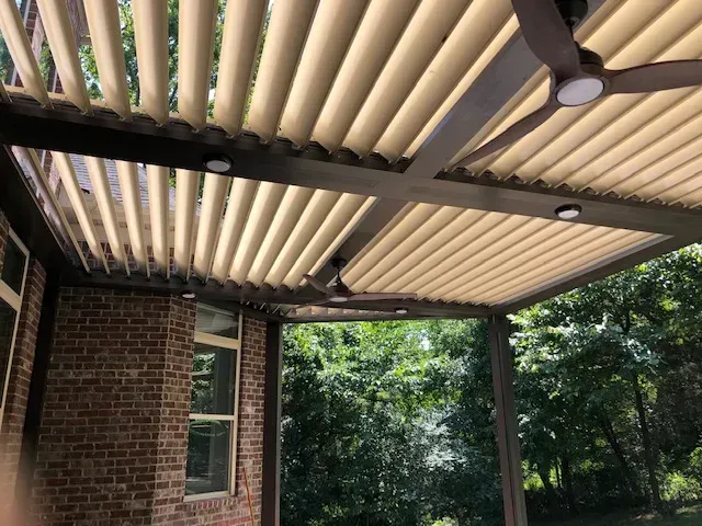 Beige louvered patio roof with brown beams, ceiling fans, and lights, attached to a brick wall.