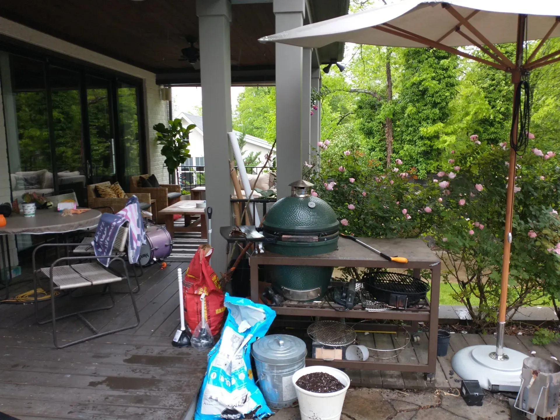 Back patio with grill, table, chairs, and umbrella. Green egg smoker on metal stand, surrounded by gardening supplies.