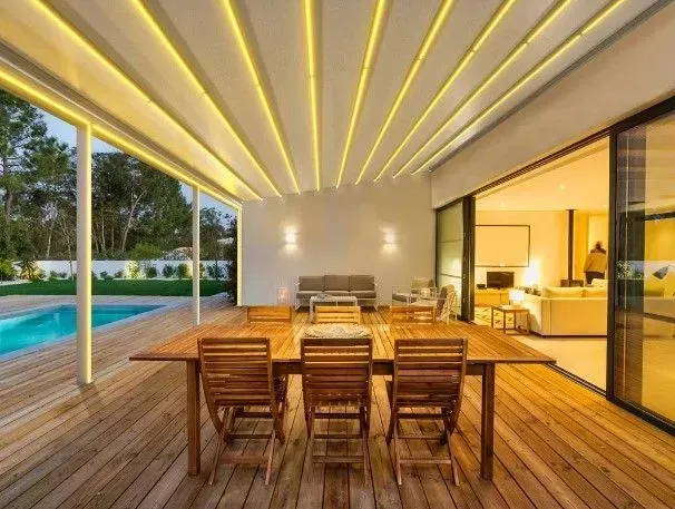Outdoor patio with wooden furniture, pool, retractable awning with yellow lights.