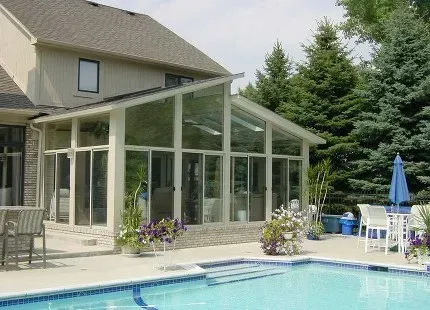 Sunroom addition with glass walls attached to a two-story beige house, overlooking a swimming pool.