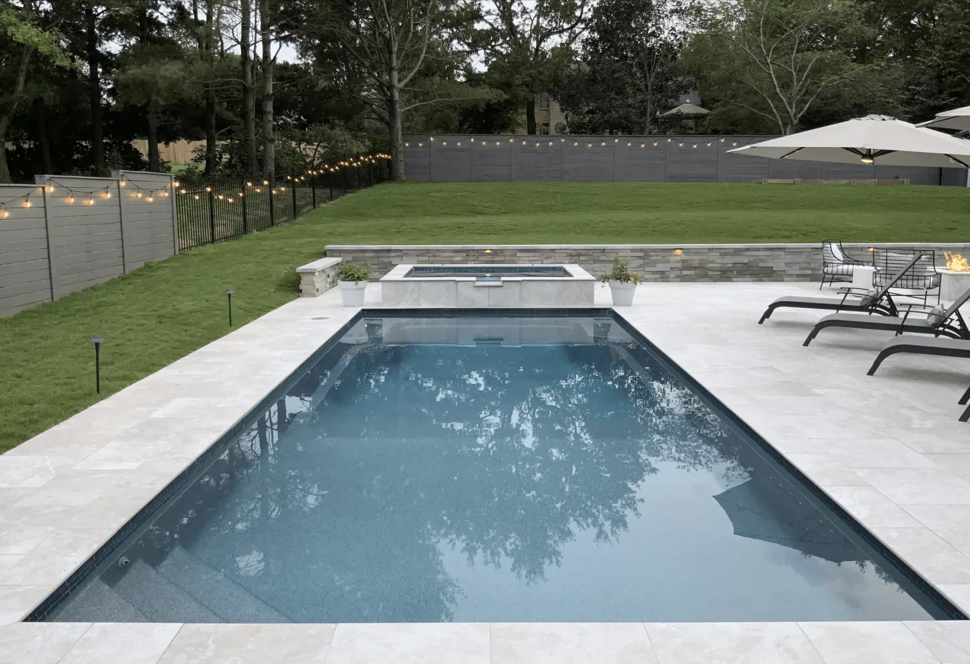 Rectangular pool with jacuzzi, lounge chairs, and manicured lawn. Gray stone patio and fence, trees in background.